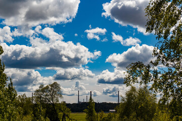 Dramatic contrast between lush green foliage and towering industrial smokestacks beneath a vibrant blue sky filled with white and grey clouds