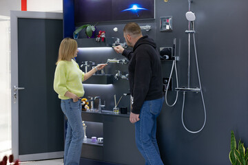 Plumbing store showroom. A consultant is explaining the features of various modern bathroom faucets and shower systems displayed on a dark wall to a male customer. Small business retail.