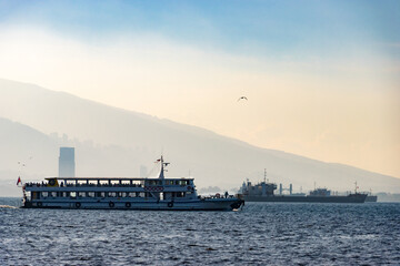 Passenger ferry crossing coastal bay with birds overhead—captured in atmospheric maritime composition featuring hazy mountain silhouettes and distant cityscape. Izmir, Turkey.