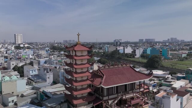 Orbit CCW drone view around Buddhist pagoda in busy urban area of Ho Chi Minh City, Vietnam on a sunny clear day featuring transportation infrastructure of main roads, canals and city skyline.