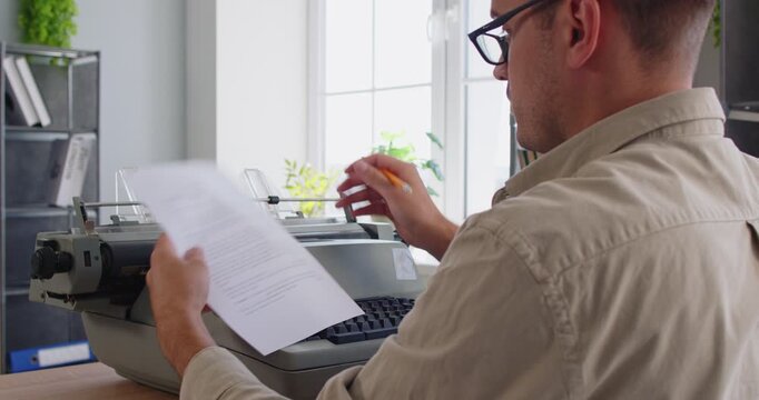 Busy editor and writer doing proofreading paper document typing on vintage typewriter. Back view of author holding manuscript and pencil to read and edit creative draft, revision before publishing