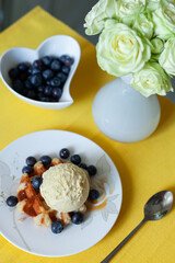 Delicate breakfast scene: waffle topped with vanilla ice cream and fresh blueberries on a floral plate. Heart-shaped bowl of blueberries, white vase with pale green roses, yellow tablecloth.