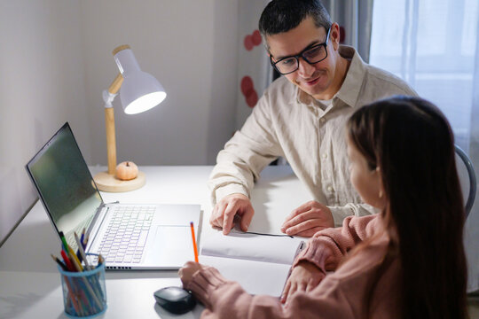 A father assists his young daughter with her homework at a brightly lit desk. They both appear engaged in the task, surrounded by school supplies and a laptop