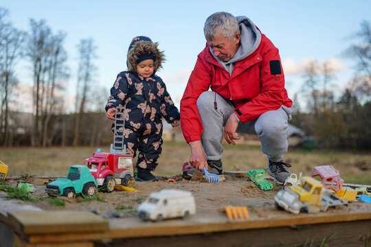 A grandfather and his young grandchild enjoy an afternoon in a sandbox, playing with various toy vehicles. The sun shines brightly in the background - Powered by Adobe