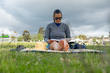 Female adult reading outdoors on blanket in park with cloudy skies.