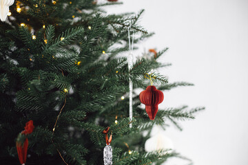 Paper red baubles on Christmas tree branches close up. Stylish holiday bauble and ornaments on decorated xmas tree with lights. Winter holidays decor, atmospheric christmas time