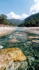 A clear river flows through a rocky canyon, surrounded by lush green vegetation and mountains under a bright blue sky with fluffy white clouds.