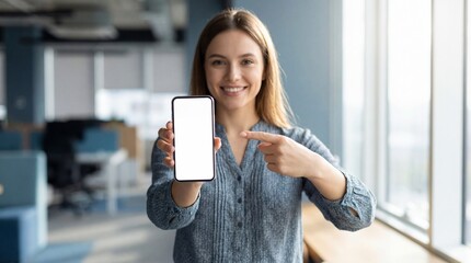 Smiling businesswoman pointing her finger at a smartphone with a blank white screen in a modern office, ideal for mockup or advertising