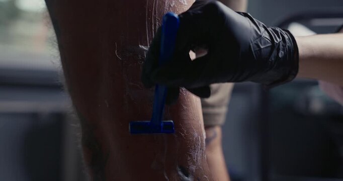 Tattoo artist in black gloves shaving a client's leg with a disposable blue razor before applying a tattoo stencil
