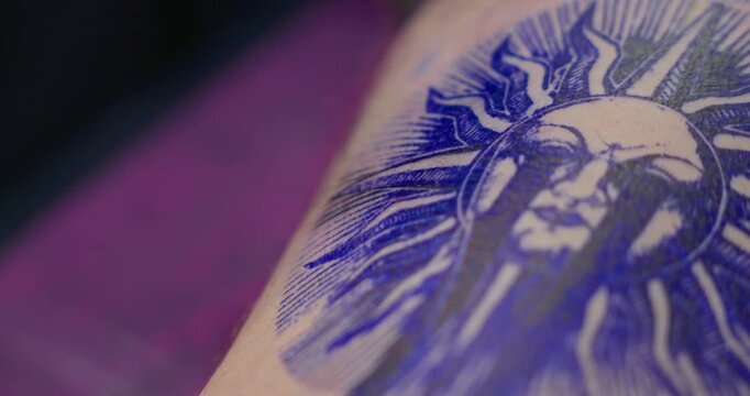 Close-up of a tattoo machine needle injecting ink into the skin, outlining a sun design on a leg