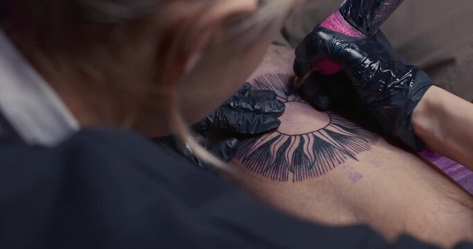 Close-up of a tattoo machine needle injecting ink into the skin, outlining a sun design on a leg
