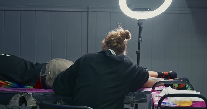 Silhouette of a female tattoo artist preparing a client for a session, standing against a window and a ring light in a studio