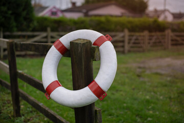 A white lifebuoy with red bands is mounted on a wooden post beside a fence in a rural grassy area, fast river flows nearby. This image conveys safety, outdoor life, and community protection.
