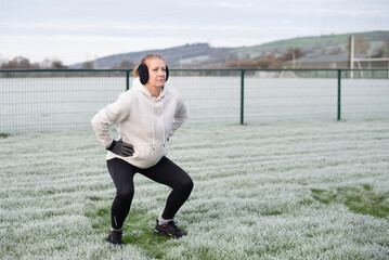 A middle aged woman in a light hoodie, gloves, and earmuffs performs a squat in frosty field. Outdoor winter fitness, workout  scene highlighting focus, energy, and athletic effort