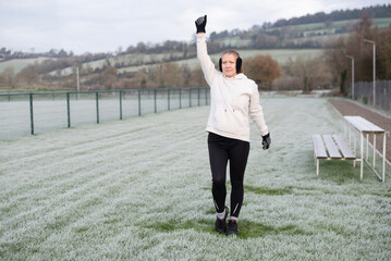 A determined woman outdoors in a white hoodie and gloves raises her fist in victory gesture on frosty field. Woman starts training outside in winter, does gymnastics, warms up before training