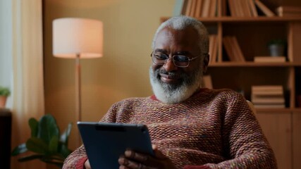 Elderly man enjoys reading on tablet in cozy living room while relaxing in comfortable chair with smile and joy - Powered by Adobe