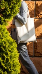 Woman in stylish grey pinstripe trousers and long-sleeve top carrying a blank cream canvas tote bag outdoors against a textured brick wall with green foliage. customizable eco-friendly mockup.