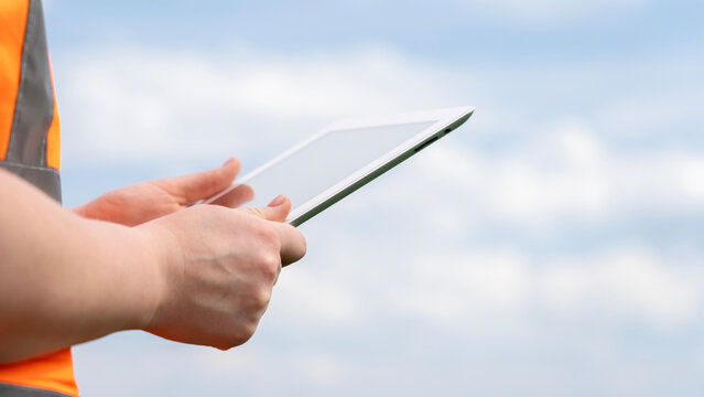 A person wearing an orange vest is holding a tablet in their hand. The sky is cloudy and the person appears to be focused on the tablet