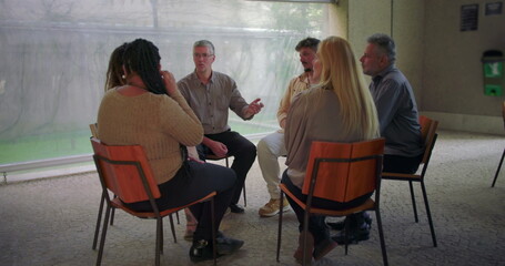 Therapy group session with diverse individuals seated in circle, participants sharing and listening during guided emotional support meeting indoors
