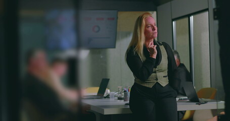 Thoughtful blonde businesswoman seated during office meeting, hand on chin while observing data screen, colleagues engaged in discussion