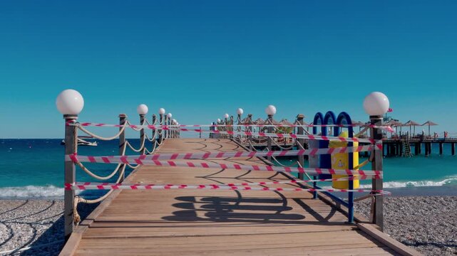 The pier is closed to vacationers due to rough seas and strong winds. Waves crash against the breakwaters, and the strips of the pier flutter and sway in the gusts of wind. The deserted shore and stor
