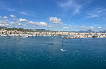 Scenic panoramic view of a coastal city with marina, yachts, and calm blue sea under a bright sky with scattered clouds, surrounded by hills and buildings