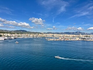 Scenic panoramic view of a coastal city with marina, yachts, and calm blue sea under a bright sky with scattered clouds, surrounded by hills and buildings