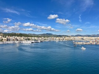 Scenic panoramic view of a coastal city with marina, yachts, and calm blue sea under a bright sky with scattered clouds, surrounded by hills and buildings