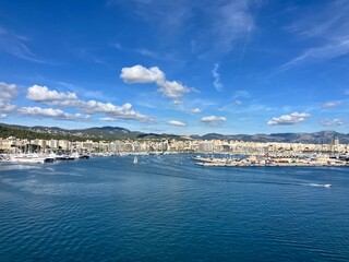 Scenic panoramic view of a coastal city with marina, yachts, and calm blue sea under a bright sky with scattered clouds, surrounded by hills and buildings