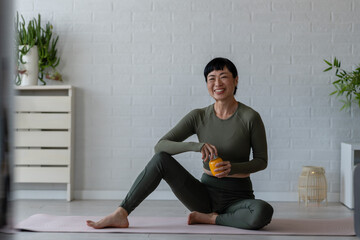 Smiling Asian woman relaxing on a yoga mat at home, holding a bottle of vitamins after a workout,...