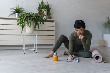Woman in sportswear sitting on floor by yoga mat, writing in notebook and reflecting after home...