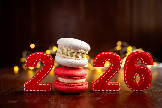 Red fabric numbers forming 2026 with a stack of delicious macarons, creating a festive New year celebration setup with blurred bokeh lights on a dark background