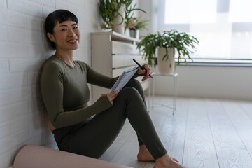 Smiling woman in sportswear sitting on floor at home with notebook, relaxing after workout and...