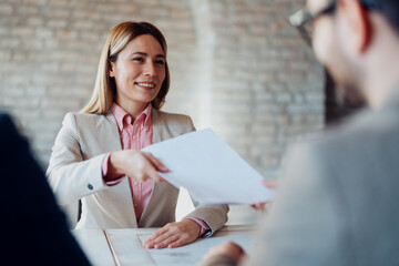 A female manager is passing a report or agreement paperwork to a male employee across a desk in a modern office setting, discussing business details.