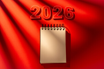 Red felt numbers 2026 and a blank spiral notebook lying on a vibrant red background, bathed in diagonal sunlight shadows, symbolizing future planning, New year resolutions, fresh starts