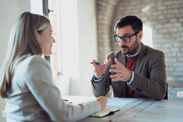 A professional man and woman sit across from each other at a table during a business meeting. The man, wearing glasses and a suit jacket, gestures with his hands while explaining a point.