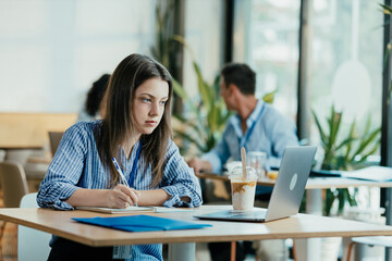 Fototapeta premium Female Student Taking Notes from Online Lecture in Sunny University Cafeteria During Lunch
