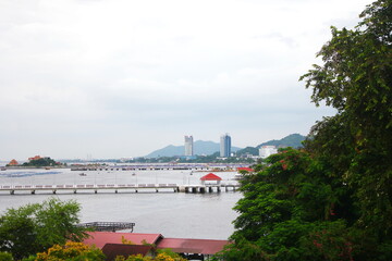 A scenic coastal view with a long pier extending into the sea, framed by lush green trees and hillside buildings under an overcast sky.