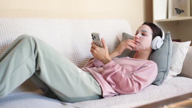 A young woman lying on a sofa using her phone while wearing a cosmetic facial mask. Concept self-care at home. - Powered by Adobe