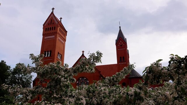 Church of Saints Simon and Helena known as Red Church in Minsk stands behind blooming branches under dramatic cloudy sky in spring, Minsk, Belarus, taken handheld. High quality 4k footage