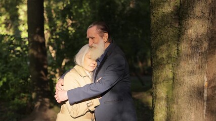 Grandfather and grandmother in love hugging outdoors in a city park near a large tree in sunlight.