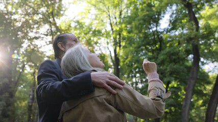 Pensioners in love stand among trees against the background of sunlight, hugging each other