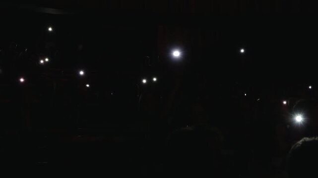 A dark scene illuminated by scattered bright phone or flashlight beams from a crowd, creating starburst effects in a dimly lit environment.