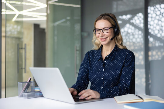 Customer service agent in a modern call center smiling and typing on a laptop while wearing a headset, offering friendly, professional technical and sales support at her desk