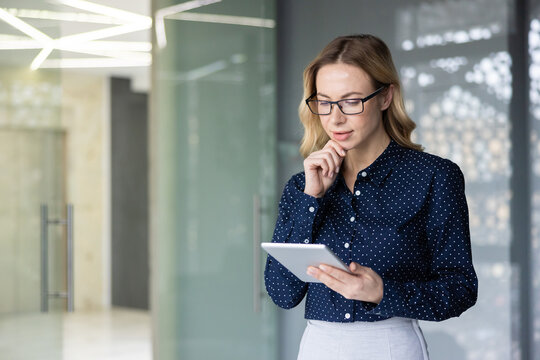 Blonde businesswoman in glasses with hand on chin is concentrating while reading information on a portable digital tablet, standing in a contemporary corporate office environment