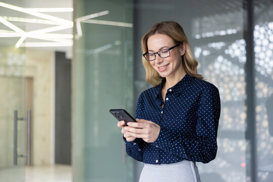 Young businesswoman smiling, holding and looking down at her smartphone while networking, communicating, and managing business tasks in a modern corporate office setting