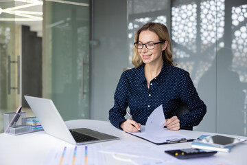 Smiling businesswoman in glasses completing financial paperwork at her modern office desk, managing documents and using a laptop for daily business operations