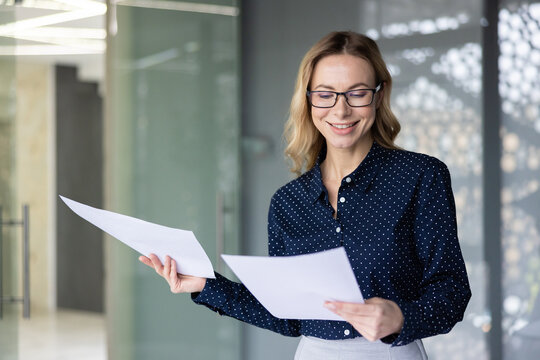 Smiling businesswoman wearing glasses standing in an office, reviewing documents and reports with a positive expression, representing success, professionalism, and achievement in her career