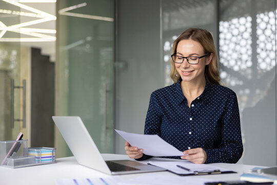 Professional businesswoman smiling while reading paperwork at a modern office desk, demonstrating job satisfaction, professionalism, and effective workflow
