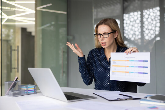 Businesswoman presenting financial metrics during a video conference, showing confusion and frustration while discussing data on her laptop in a modern office